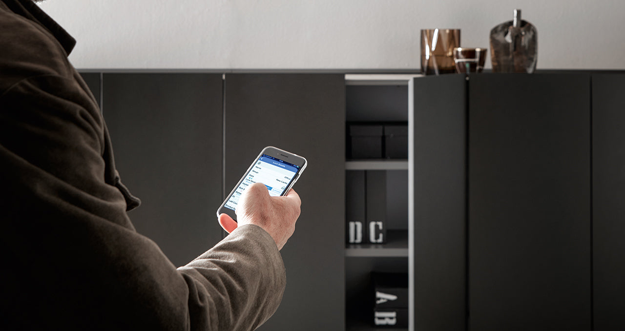 Une personne utilise un smartphone devant les armoires sombres Kyo d'Attitudes Bureaux, qui présentent des matériaux haut de gamme, des objets de décoration et des boîtes de rangement à l'intérieur et à l'extérieur des étagères.