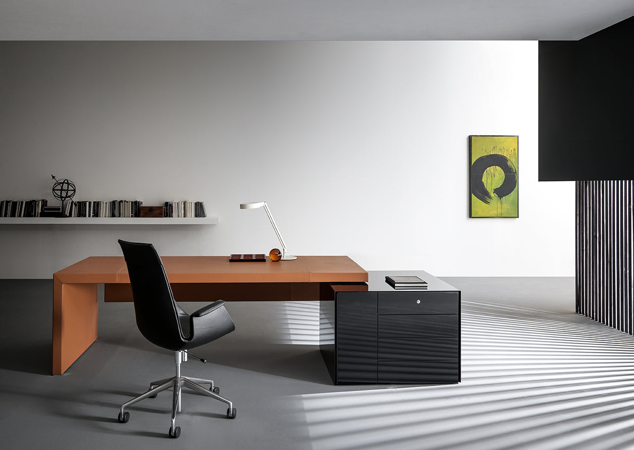 Un bureau minimaliste présente le bureau Kyo d'Attitudes Bureaux - grand, marron et fabriqué à partir de matériaux de première qualité - associé à une chaise pivotante noire, une armoire noire, une lampe de bureau, des livres sur une étagère, des œuvres d'art vertes et la lumière du soleil sur le sol gris.