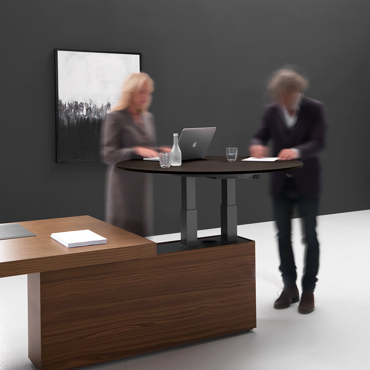 Deux personnes se tiennent debout devant une table de réunion ronde Kyo moderne et réglable d'Attitudes Bureaux, fixée à un bureau en bois. Un ordinateur portable, des verres et une carafe d'eau sont posés sur la table ; un tableau noir et blanc est accroché au mur sombre situé derrière.