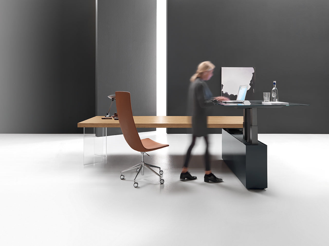 Une femme tape sur le bureau assis-debout Kyo d'Attitudes Bureaux, fabriqué à partir de matériaux haut de gamme, dans un bureau minimaliste avec une chaise brune, une lampe, une bouteille d'eau, des murs gris et une lumière vive.