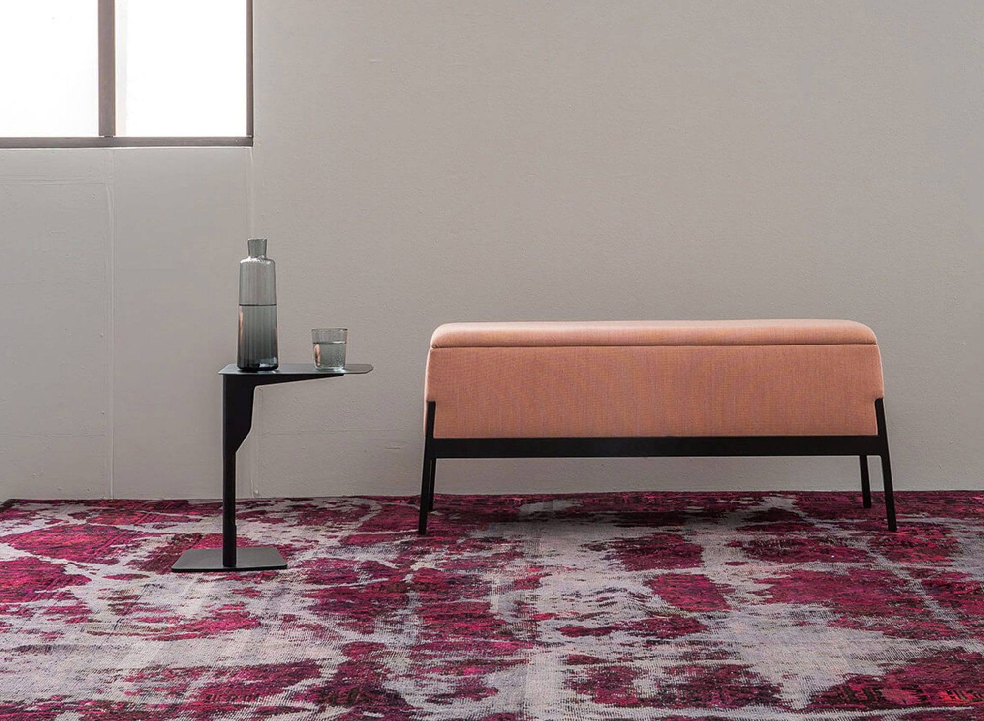 Une pièce minimaliste présente une banquette capitonnée rose, une table en métal Luca Nichetto exposant une bouteille en verre et un verre à boire, ainsi qu'un tapis à motifs rouges et gris, le tout placé contre un mur blanc uni avec une fenêtre.