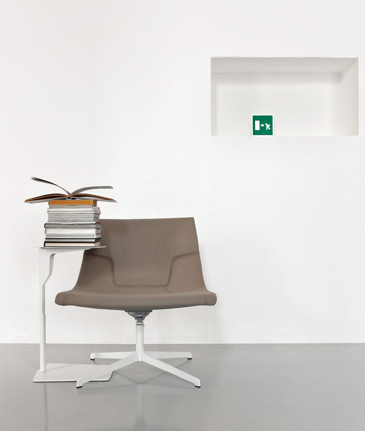 Une chaise brune moderne est assise à côté d'une table en métal épurée de Luca Nichetto, empilée avec des livres, l'un d'entre eux étant ouvert. L'arrière-plan est constitué d'un mur blanc uni et d'une étagère encastrée affichant un panneau vert de sortie de secours.