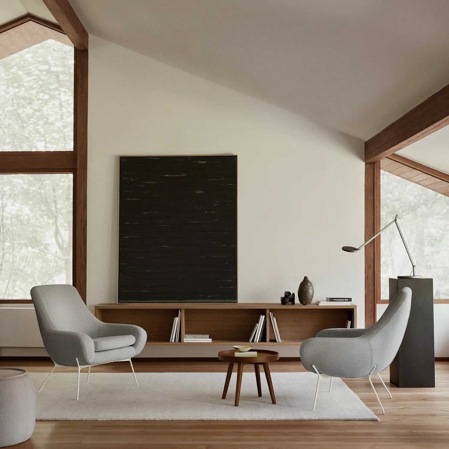 Salon minimaliste avec deux fauteuils gris modernes, dont le Fauteuil NOOMI de Susanne Grønlund avec base graphique, une table basse ronde, un tapis blanc et une console en bois. Les grandes fenêtres et le plafond en pente offrent une lumière naturelle abondante.
