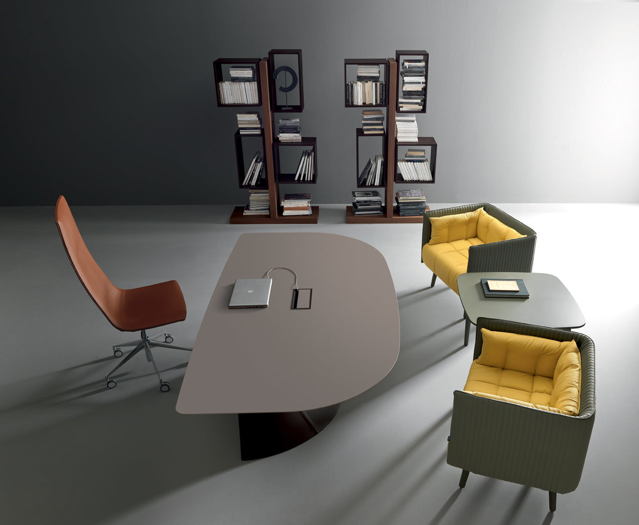 Un bureau moderne est composé d'un bureau Attitudes Bureaux Ola avec une base en ellipse, d'une chaise pivotante marron, de deux fauteuils à coussin jaune, d'une table d'appoint et de deux hautes étagères remplies de livres contre un mur gris.