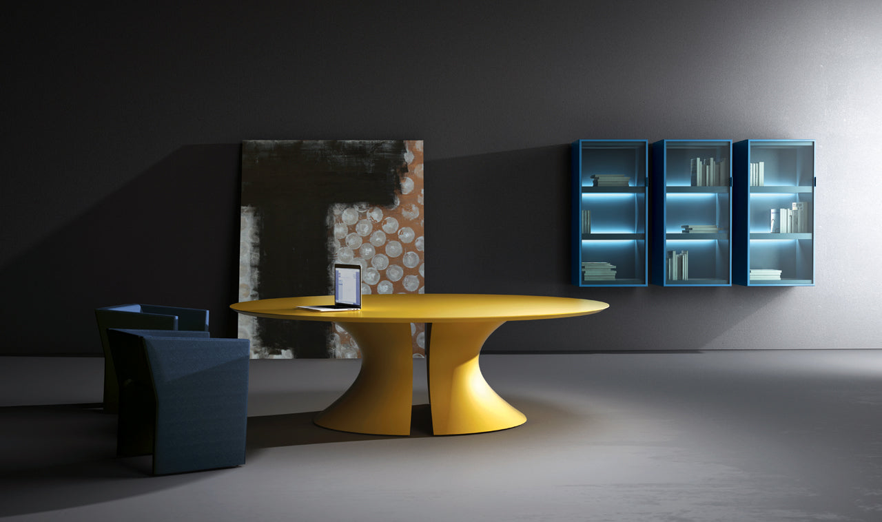 Une pièce minimaliste avec Ola, une table ronde moderne jaune (Attitudes Bureaux) avec un piètement en ellipse, deux chaises bleu foncé, un grand tableau abstrait au sol et trois bibliothèques murales bleues éclairées doucement.