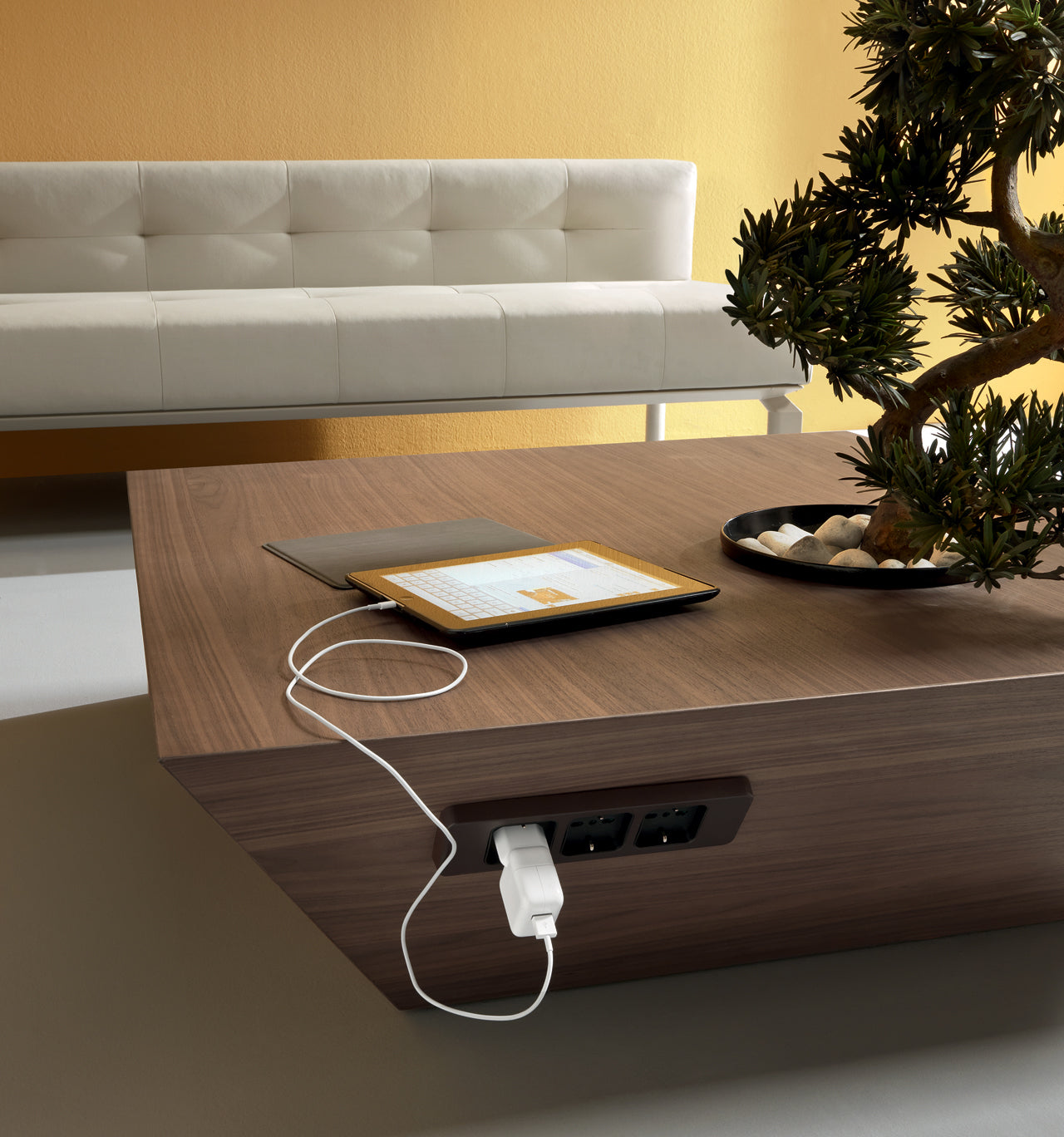 Un salon moderne avec la table basse Ola d'Attitudes Bureaux, dotée d'une base elliptique en bois et de prises électriques intégrées, d'une tablette de recharge, d'une plante décorative et d'un canapé blanc contre un mur jaune.