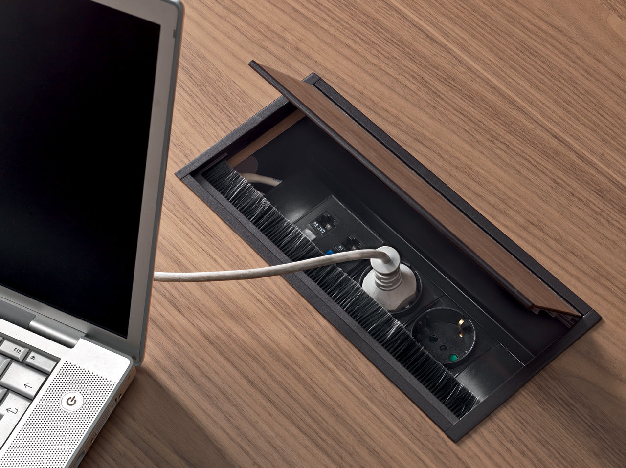 Un ordinateur portable argenté est branché sur le panneau de prises de courant Ola d'Attitudes Bureaux, intégré dans un bureau en bois au design sculptural. Le panneau partiellement ouvert révèle des prises et un câble connecté.