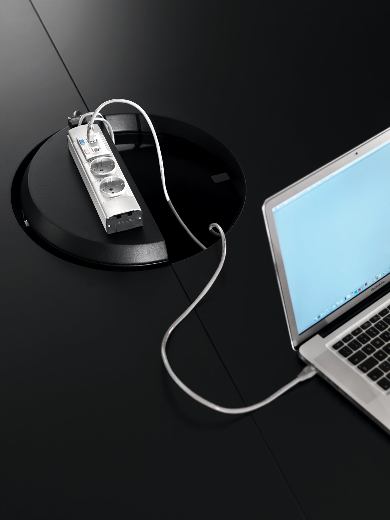 L'Attitudes Bureaux Ola, un ordinateur portable argenté avec un écran bleu, est relié à une multiprise dans un compartiment noir circulaire sur une table noire élégante avec une base elliptique, ce qui ajoute une touche de design sculptural.