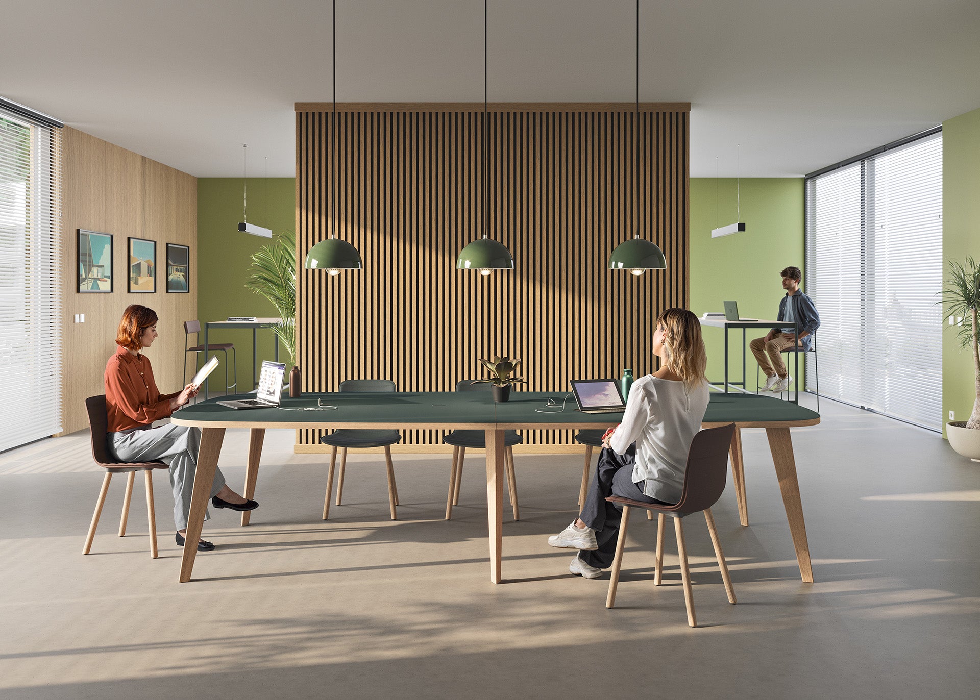 Trois personnes travaillent dans un bureau moderne aménagé avec du mobilier de direction Pigreco par Attitudes Bureaux - murs verts, accents en bois, grandes fenêtres. Deux d'entre elles utilisent des postes opératifs avec des ordinateurs portables ; l'une d'entre elles se tient debout à un bureau élevé à l'arrière-plan.