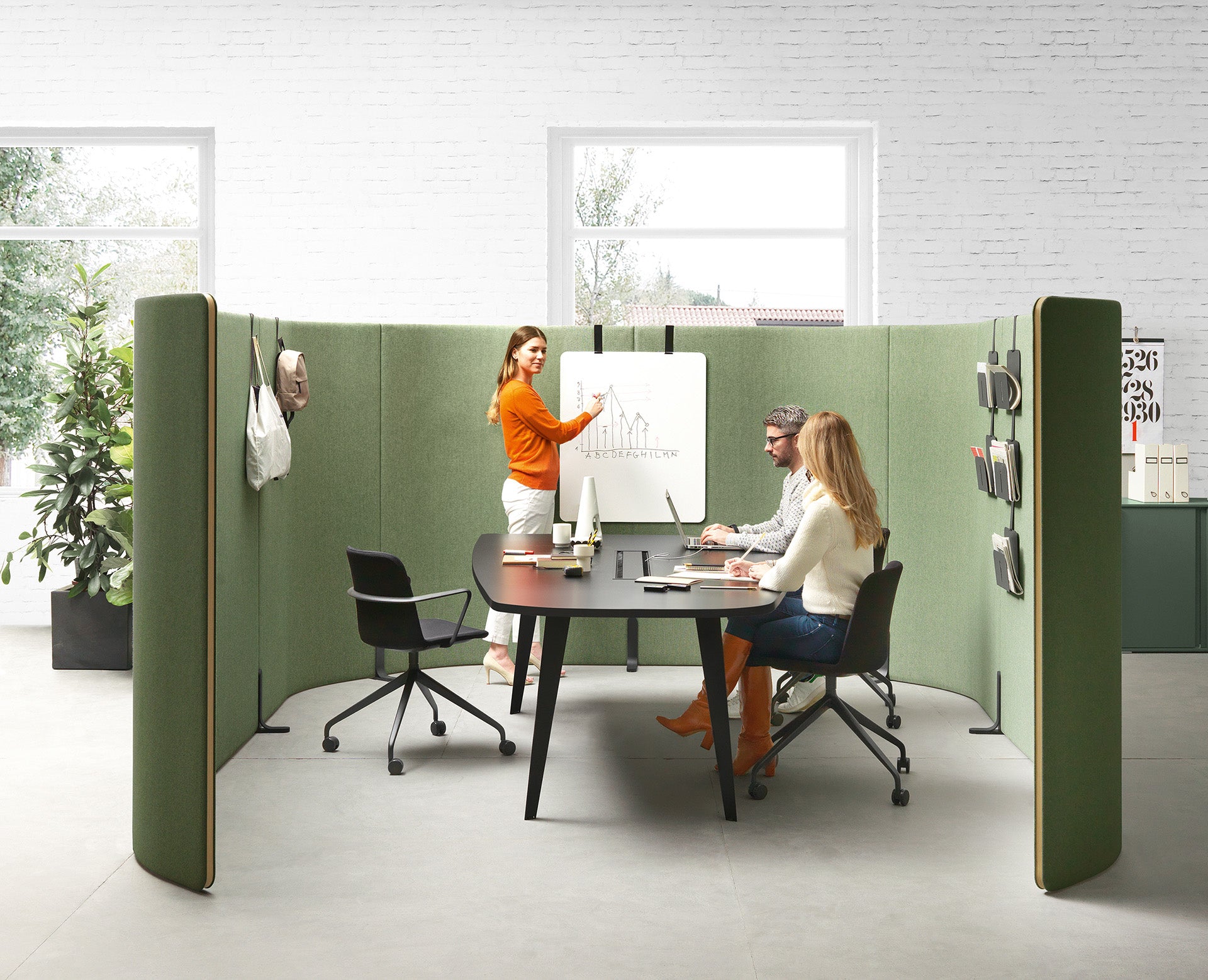 Trois personnes collaborent dans une salle de réunion moderne meublée avec Mobilier de direction Pigreco par Attitudes Bureaux. Une femme présente un graphique sur un tableau blanc, tandis que deux autres personnes travaillent à une table avec des ordinateurs portables et des cahiers.