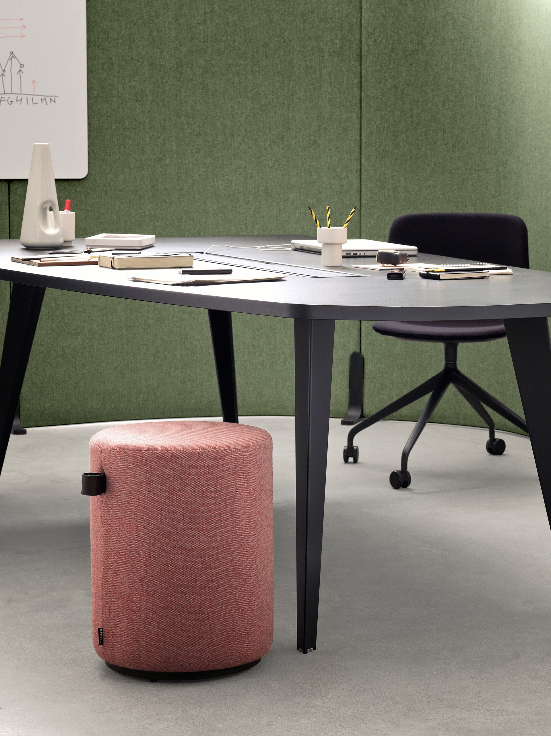 Un bureau moderne présente le Mobilier de direction Pigreco d'Attitudes Bureaux, avec un bureau noir, une chaise noire à roulettes, un tabouret cylindrique rose et des fournitures. L'arrière-plan est composé de panneaux muraux en tissu vert et d'un tableau blanc avec des dessins et des notes.