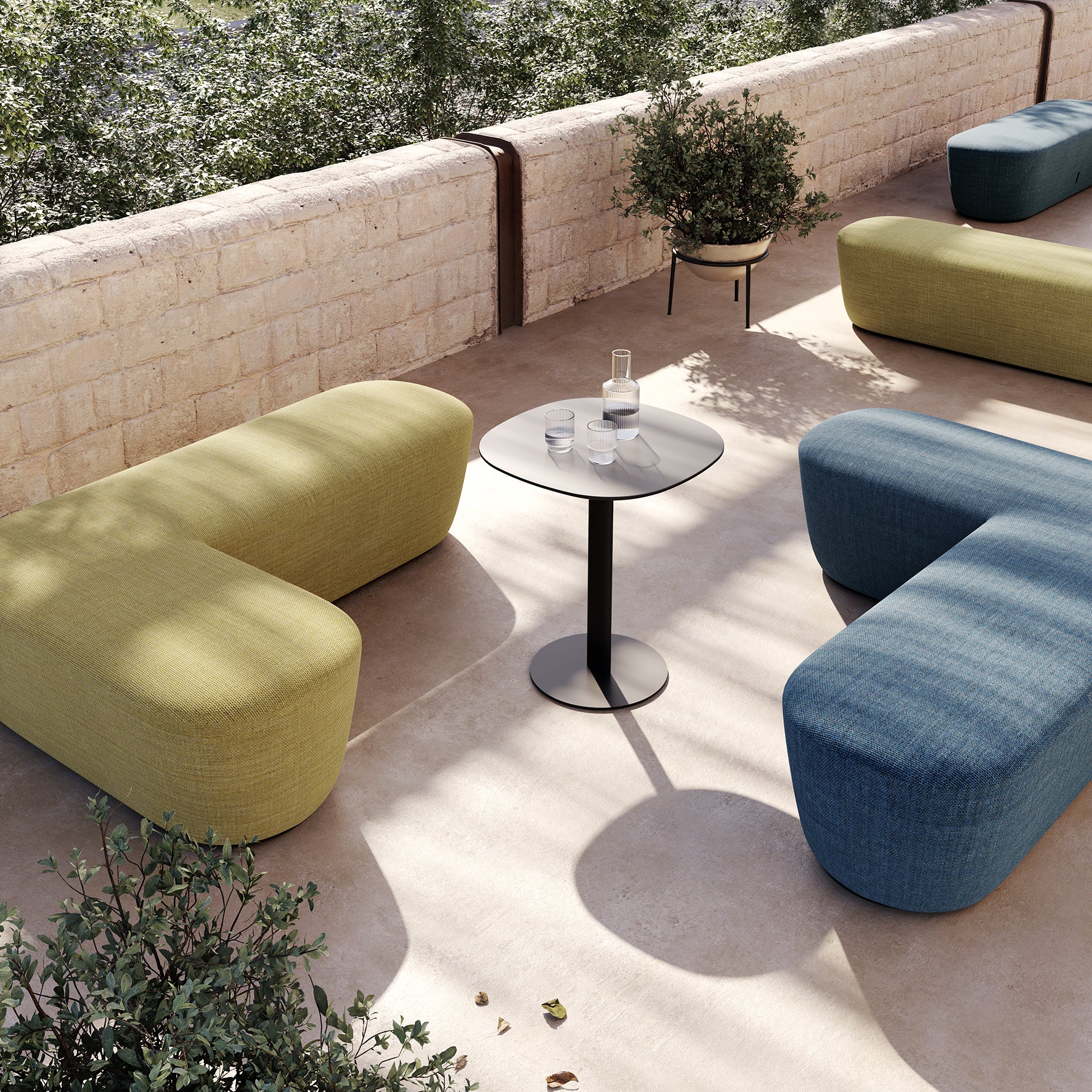 Un espace extérieur moderne comprend des bancs courbes verts et bleus de Claesson Koivisto Rune CKR qui entourent une table Tables basses Radar équipée d'une carafe et de verres. Encadrée par des murs en pierre et de la verdure, la lumière du soleil projette des ombres douces sur le sol carrelé.