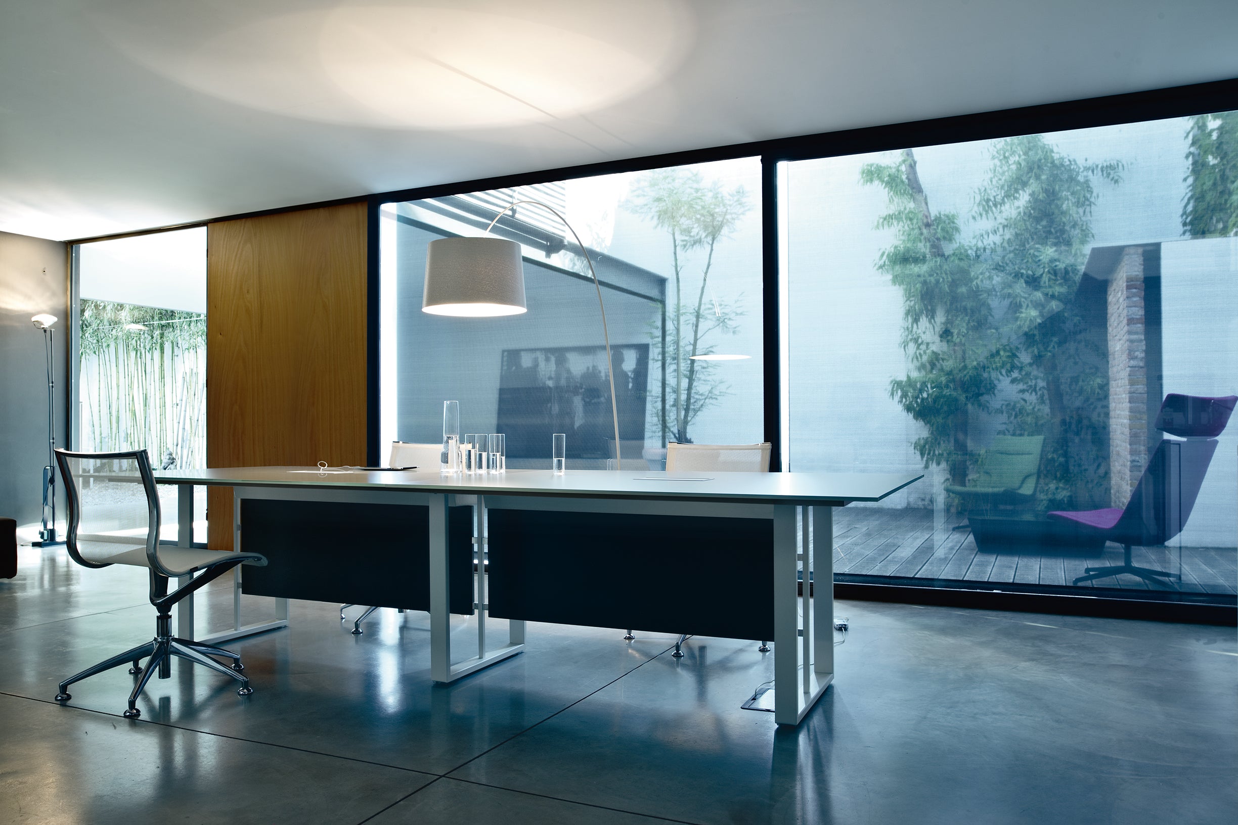 Un bureau moderne où l'on retrouve les Attitudes Bureaux Tables de réunion Velvet, de la verrerie, une chaise en maille et un grand lampadaire. Les grandes baies vitrées donnent sur la verdure et les sols en béton lisse avec connectivité intégrée conviennent aux espaces de travail contemporains.