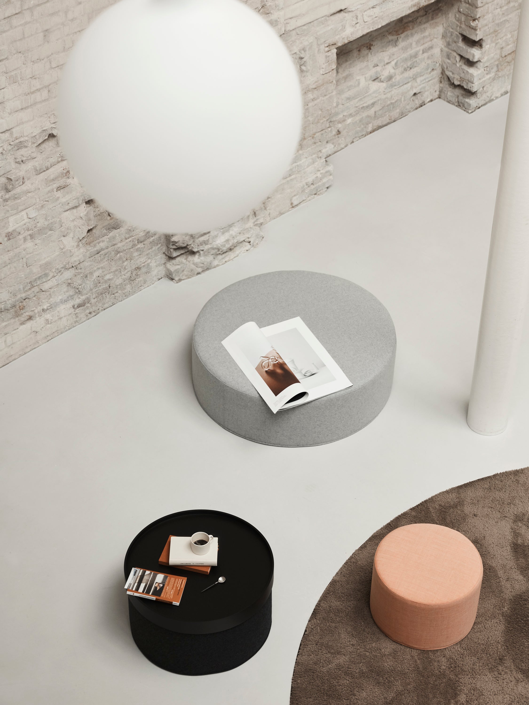 Dans une pièce minimaliste, le pouf DRUM multifonction d'Attitudes Bureaux est associé à un ottoman rond gris avec un magazine ouvert, une table basse ronde noire avec une tasse et un livret, un ottoman pêche, un tapis marron, un mur en briques apparentes et une grande lampe blanche suspendue.