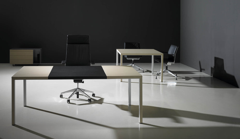 Un bureau minimaliste composé de trois bureaux Neta Executive d'Attitudes Bureaux, de deux chaises noires et d'un petit meuble à tiroirs beige sur un mur gris foncé et un sol blanc brillant. Un éclairage puissant crée des ombres marquées, ce qui est idéal pour les espaces de travail modernes.