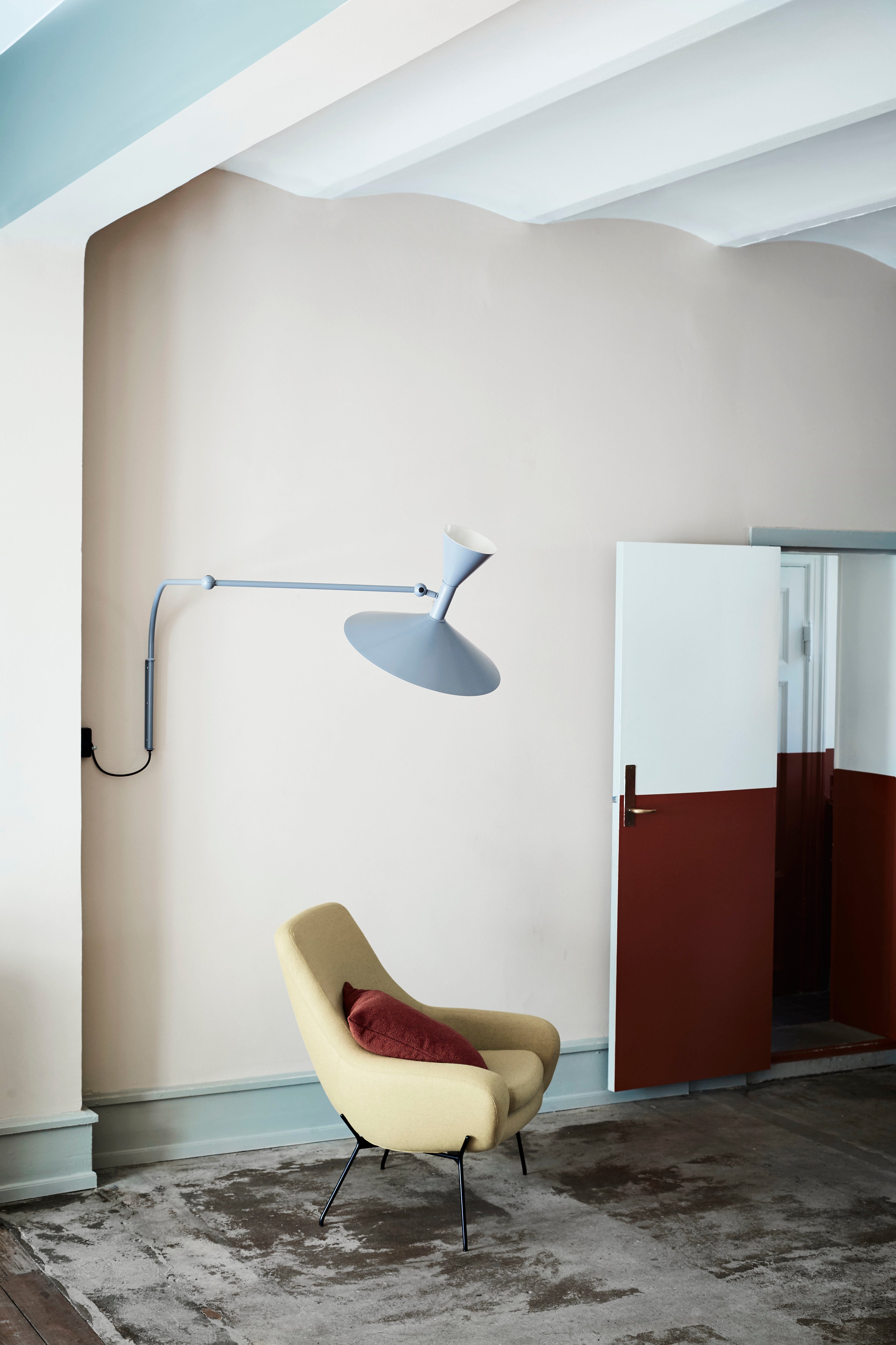 Une pièce moderne avec un mur beige clair, une lampe murale bleue à piètement graphique, un Fauteuil NOOMI de Susanne Grønlund en jaune avec un oreiller brun-rouge, une porte blanche et brune partiellement ouverte, et un tapis gris au sol.